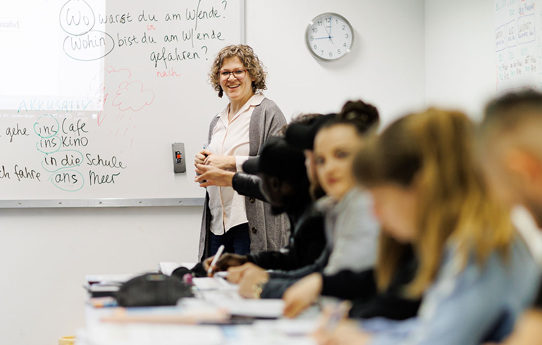 Eine Lehrerin steht am Whiteboard auf dem Notizen zu sehen sind und schaut in Ihre Klasse. Die Klasse schreibt mit.