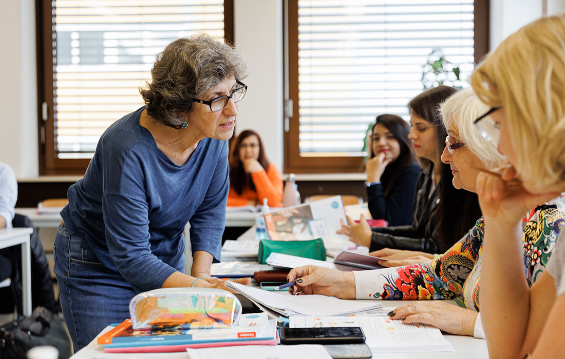 Eine Lehrerin steht auf der anderen Seite des Tisches und spricht mit einer ihrer Schülerinnen über die Aufgabe. Die Schülerinnen daneben hören gespannt zu.
