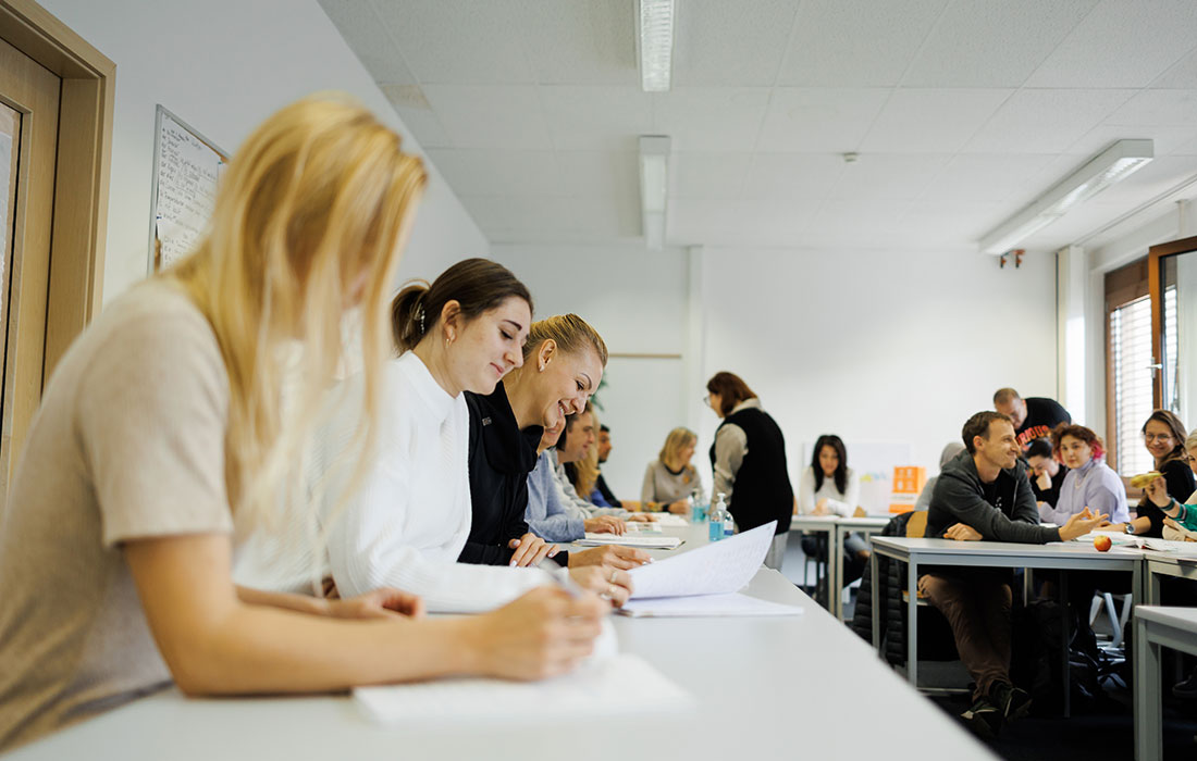 Bildausschnitt eines Klassenraums mit Sprachschülern, die gerade Gruppenarbeiten machen. Eine Lehrerin unterstützt einzelne Schüler.