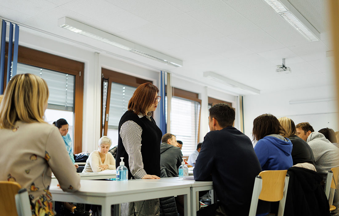 Eine Lehrerin steht in der Mitte eines Klassenraums und spricht mit ihren Sprachschülern die drumherum an Tischen sitzen und zuhören.